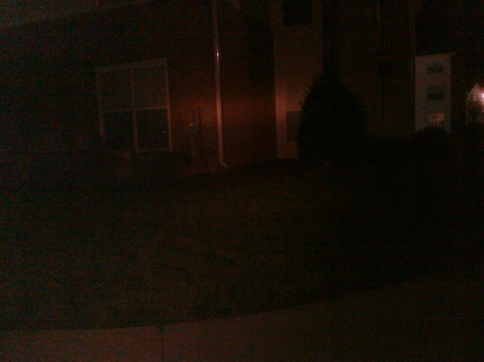 dark outdoor scene at night, shows a building with a window and a tree