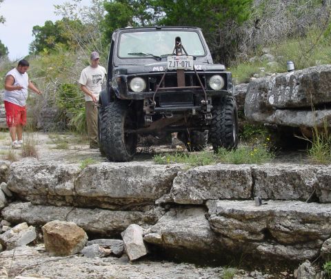 1973 Ford Bronco - Two men stand near a black off-road SUV navigating a rocky trail, suggesting an outdoor adventure or off-roading event.