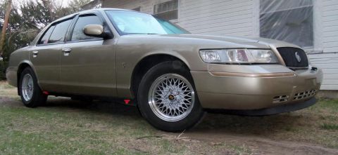 2000 Mercury Grand Marquis - A gold sedan with aftermarket wheels is parked on grass in front of a building.