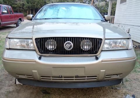 2000 Mercury Grand Marquis - Front view of a gold 2000 Mercury Grand Marquis, showing the grille and headlights. Another vehicle is visible in the background.