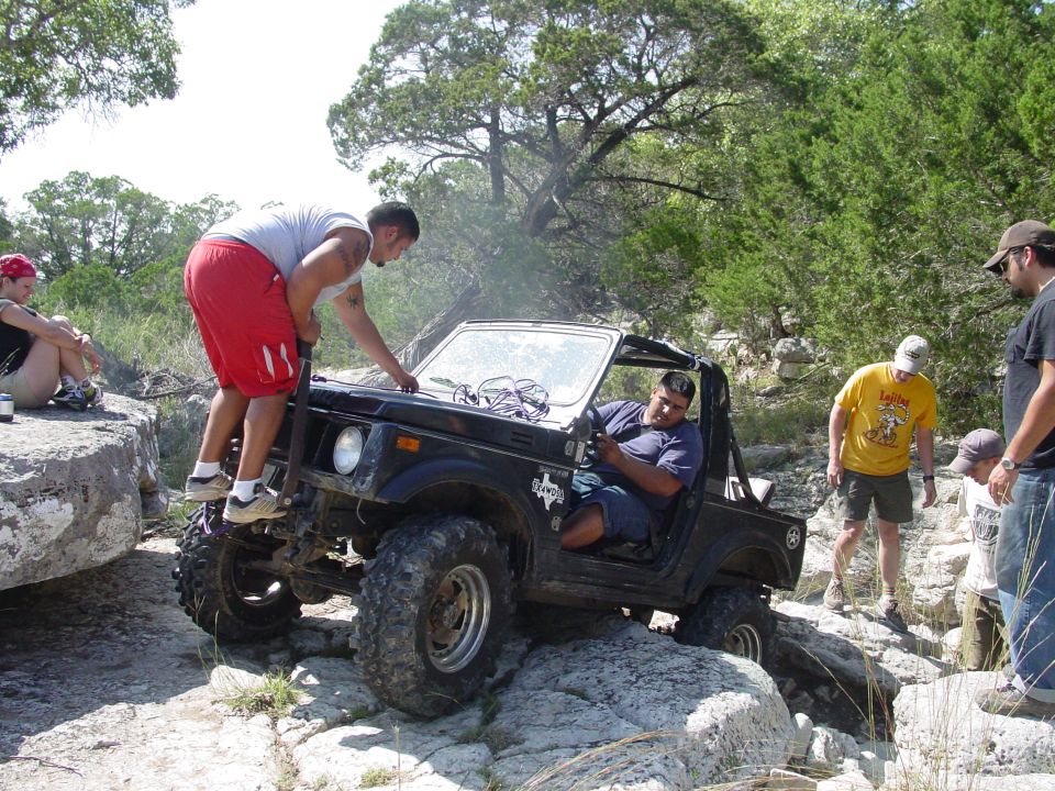Black off-road vehicle with large tires navigating rocky terrain, people around it