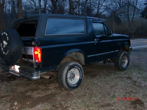 1993 Ford Bronco - A dark blue Ford Bronco is parked on a dirt lot with a house and trees in the background.