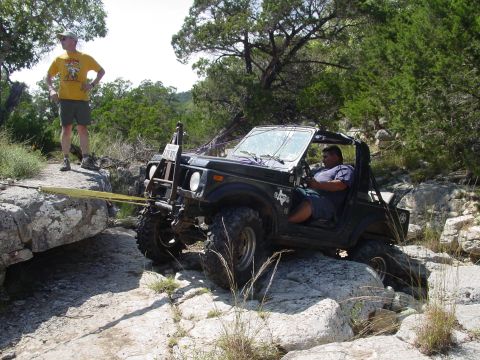 1973 Ford Bronco - A man drives an off-road vehicle over rocky terrain, assisted by a tow strap.
