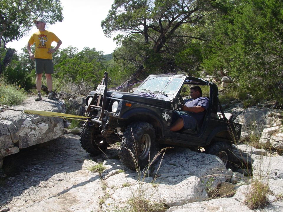 Black off-road vehicle with a man driving, on rocks, with a yellow tow strap attached to a large rock