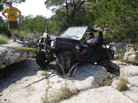 1973 Ford Bronco - A black off-road vehicle navigates a rocky trail, with a winch line secured to a large boulder.