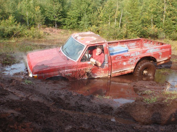 Red pickup truck stuck in mud, driver visible, trees in background