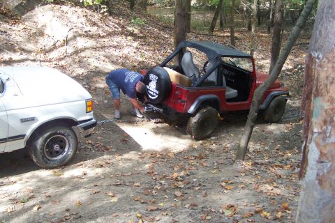 1989 Ford Bronco - A man connects a winch cable between a white Ford pickup and a red Jeep Wrangler in a wooded area.