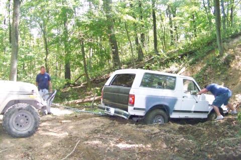1989 Ford Bronco - A white Ford Bronco is stuck in the mud while a Jeep attempts to pull it out, with a person pushing from behind.