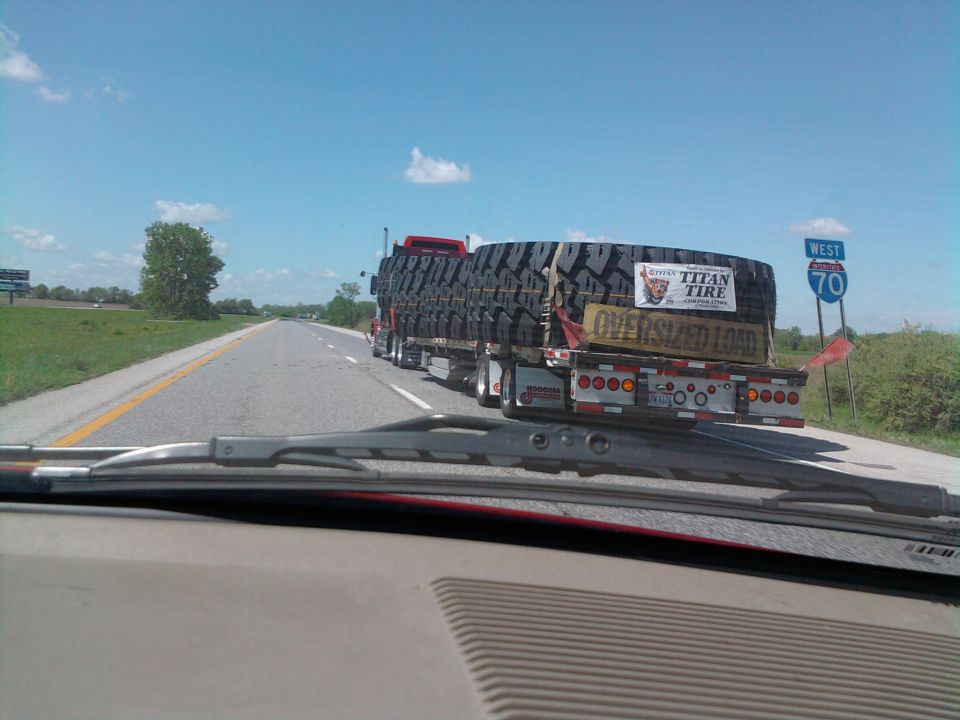 Truck trailer carrying oversized tires, with 'Titan Tire' and 'Oversized Load' signs, on a highway