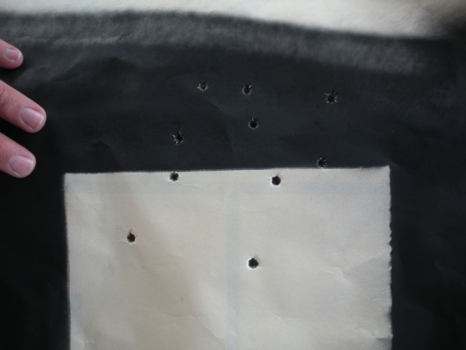 Paper target with multiple bullet holes, one corner cut out. Hand visible on left