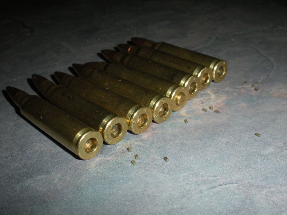 Row of brass cartridges on a gray surface with small debris scattered around. A Supermotors.net logo is in the lower right corner