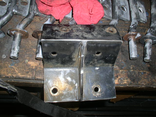 Fabricated bare-steel welded bracket with drilled holes on a workbench, manifolds behind