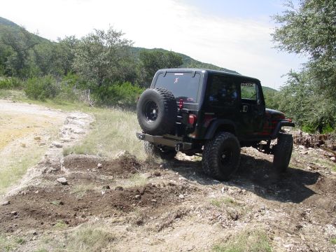 1973 Ford Bronco - A black Jeep Wrangler TJ navigates a rough, off-road trail under a partly cloudy sky.