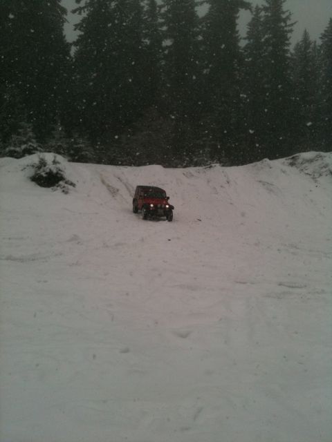 2005 Jeep Wrangler - A red 2005 Jeep Wrangler navigates a snow-covered hill amidst falling snow, with a dense forest in the background.