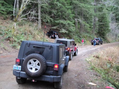 2005 Jeep Wrangler - A line of Jeep Wranglers drives down a dirt road in a wooded area. The lead Jeep is black with its spare tire mounted on the rear.