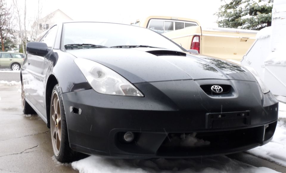 Black Toyota Celica coupe front three-quarter with matte black bumper and bronze wheels in snow