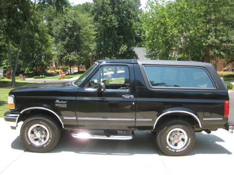 1992 Ford Bronco - A black 1992 Ford Bronco SUV is shown from the side, parked on a paved surface with trees and houses in the background.