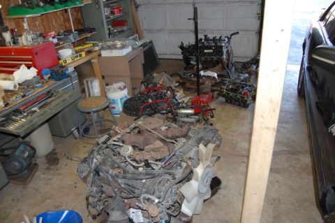 1985 Mercury Grand Marquis - Disassembled engines and automotive parts are stored in a cluttered garage next to a black vehicle.