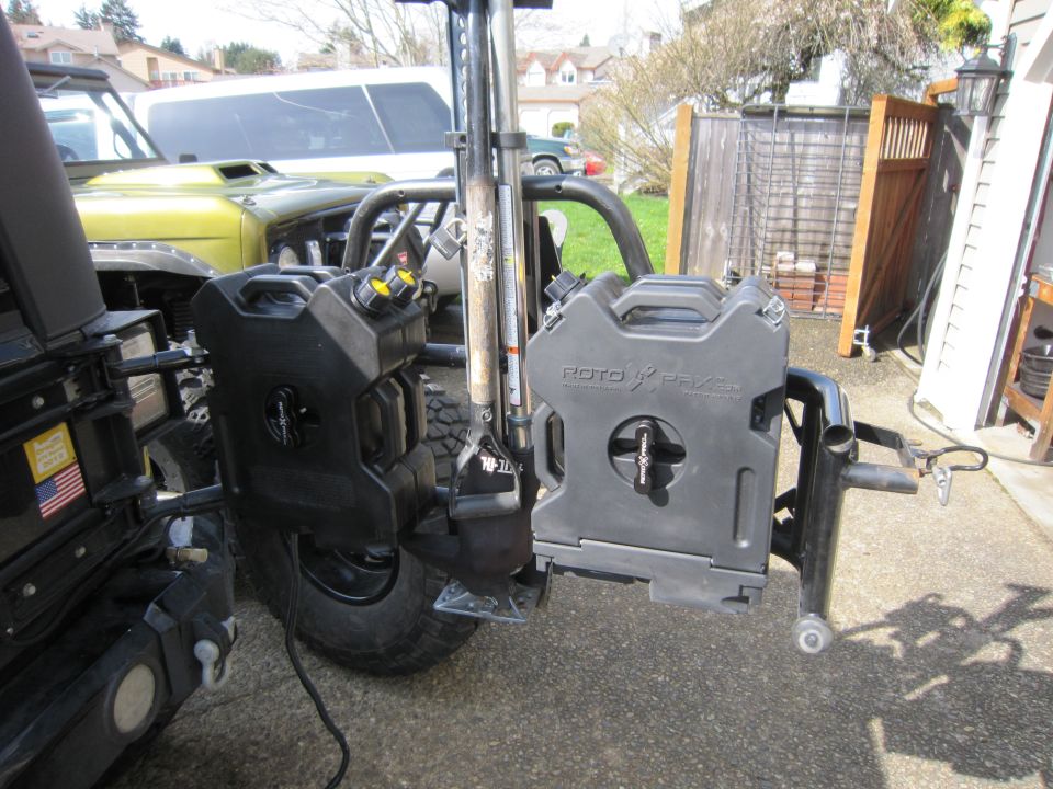 Rotopax fuel cans mounted on the rear of a dark-colored Jeep Wrangler, with a yellow Jeep visible in the background