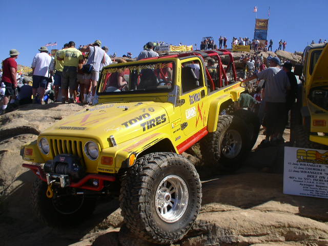 Yellow Jeep Wrangler with 'Toyo Tires' on hood, climbing rocks with crowd watching. Another Jeep is parked to the right