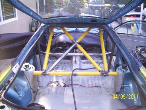 1995 Ford Probe - The interior of a car has been stripped and fitted with a yellow roll cage, with other vehicles visible in the background.