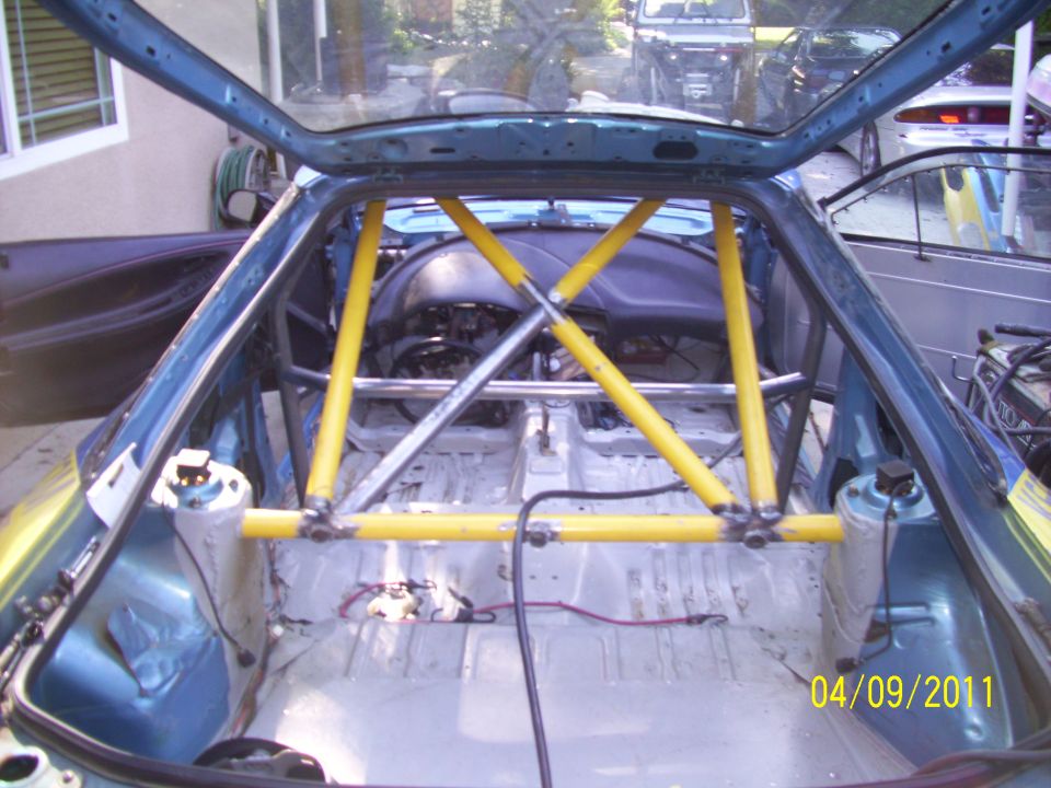 car interior stripped down with yellow roll cage installed, view from rear hatch. Other vehicles visible outside