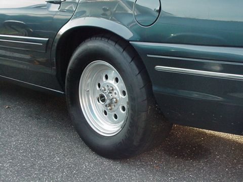 1998 Ford Crown Victoria - polished aluminum slotted mag wheel fitted with a drag-style tire on a dark green car, parked on asphalt.