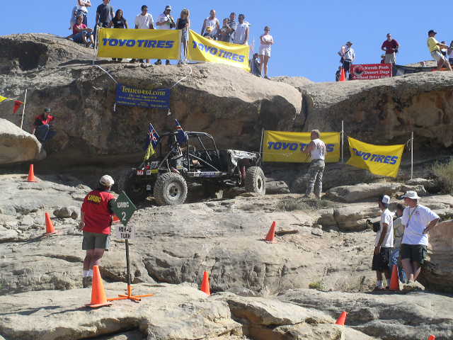 Off-road buggy with flags on rocky terrain, spectators on hill