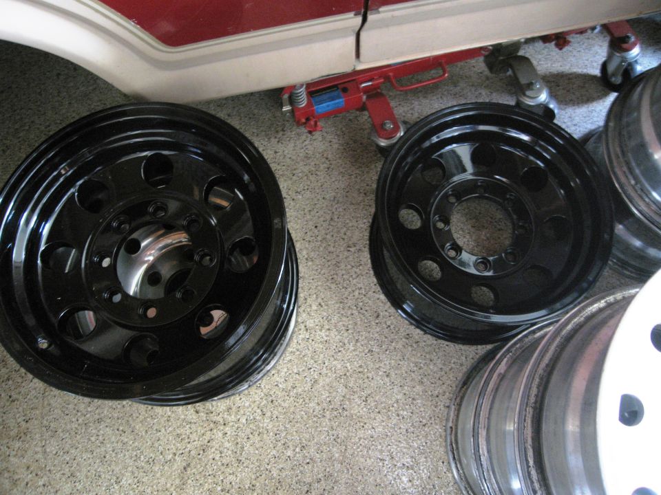 stack of black off-road wheels, one is a spare, with a red jack under a vehicle in the background