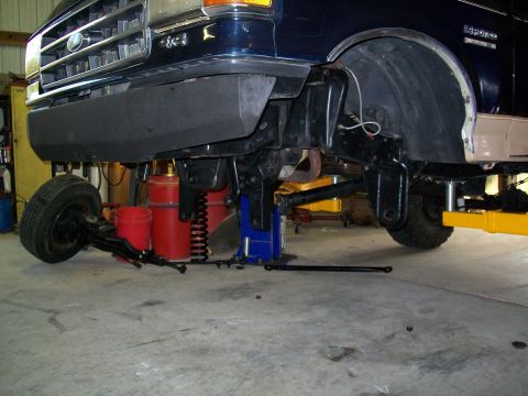1990 Ford Bronco - The front end of a dark blue Ford truck is shown lifted on a hoist, with the front wheel removed for service.