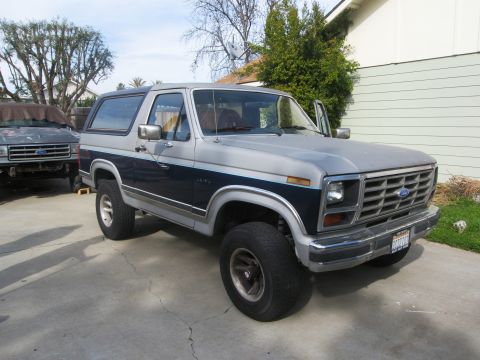 1986 Ford Bronco - A two-tone Ford Bronco is parked in front of a house, with another vehicle in the background.