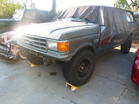1986 Ford Bronco - A gray Ford Bronco is partially covered by a tarp, parked next to another vehicle.