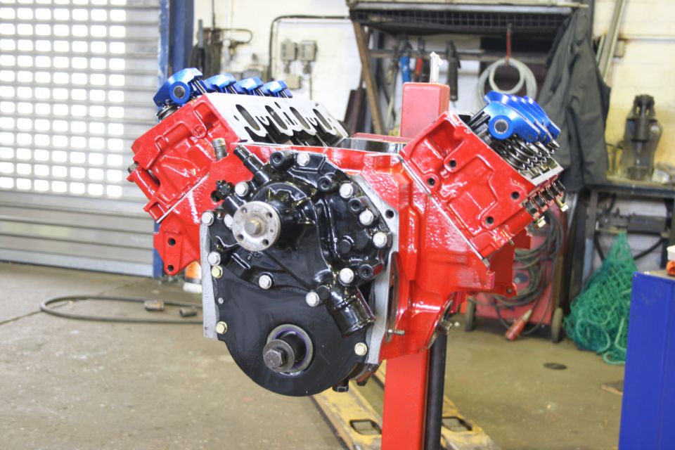 Red Ford V8 engine block with black oil pan and blue valve covers, mounted on a red stand