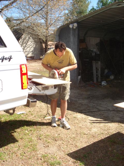 1992 Ford C-350 (Centurion Conversions) - Man uses a circular saw to cut wood resting on the tailgate of a white pickup truck.
