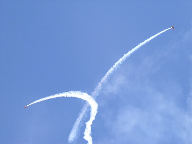 Two red biplanes trail white smoke against a blue sky, one ascending sharply, the other in a curved dive