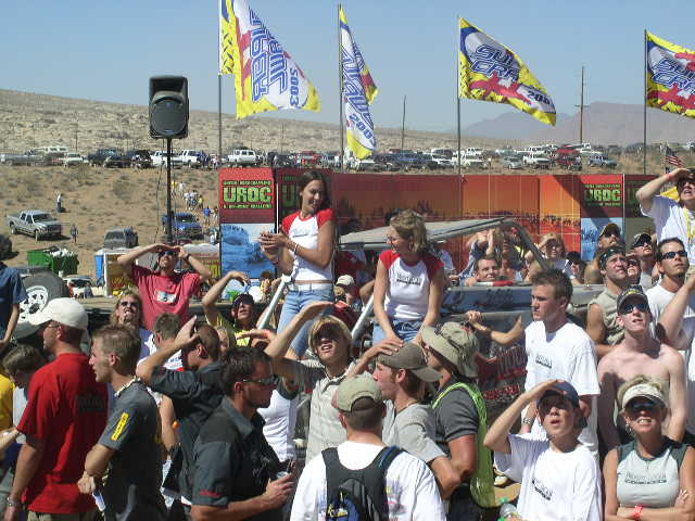 Crowd at an outdoor event with flags and parked vehicles