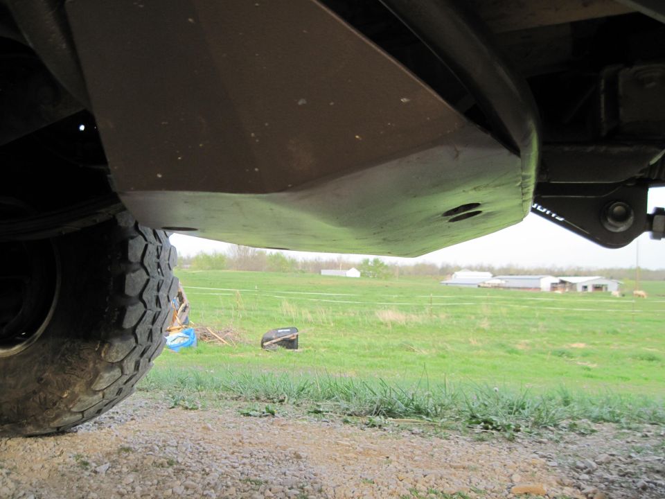 Underside view of a vehicle's skid plate and tire, with a grassy field and buildings in the background