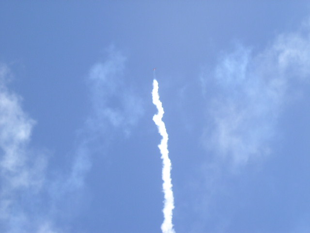 Rocket launch trail against a blue sky with clouds. Supermotors.net logo in lower right