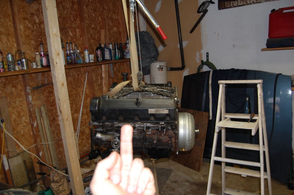 Engine assembly suspended from a hoist in a garage, with shelves of bottles in the background
