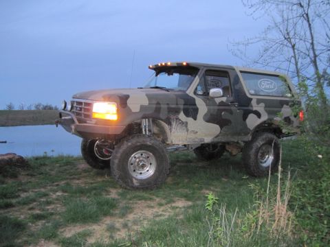 1995 Ford Bronco - This lifted Ford Bronco features a camouflage paint job and aftermarket wheels, parked by a body of water.
