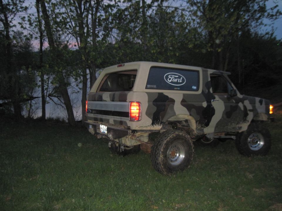 Camouflaged Ford Bronco parked on grass near water, rear view