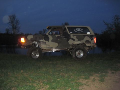 1995 Ford Bronco - A camouflaged Ford Bronco sits on a grassy embankment near water under a dark sky.
