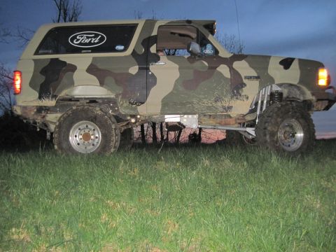 1995 Ford Bronco - Camouflaged 1995 Ford Bronco with lifted suspension and off-road tires sits in a grassy field at dusk.