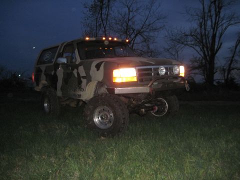 1995 Ford Bronco - A camouflaged Ford Bronco is parked on a grassy field at dusk, illuminated by its headlights and roof lights.
