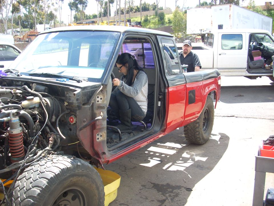 Red and black pickup truck with front end removed, woman inside, man standing behind. Another truck is parked nearby