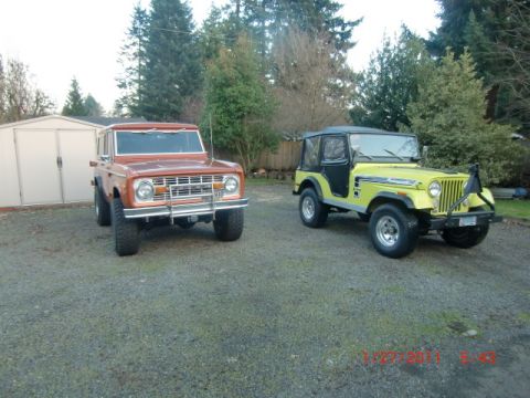 1973 Ford Bronco - Two classic off-road vehicles, a 1973 Ford Bronco and a Jeep CJ-5, are parked outside on a gravel driveway.