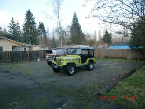 1973 Ford Bronco - Two classic off-road vehicles are parked in a gravel lot with houses and trees in the background.