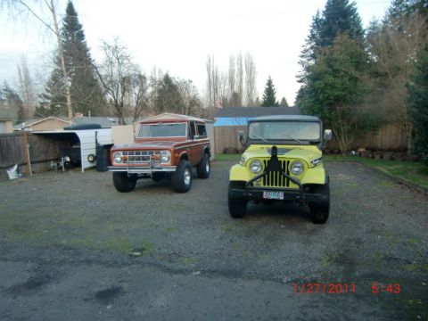 1973 Ford Bronco - A brown 1973 Ford Bronco and a yellow Jeep are parked side-by-side on a gravel lot with trees and buildings in the background.