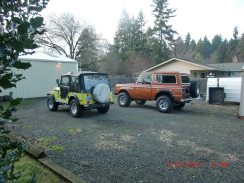 1973 Ford Bronco - Two classic off-road vehicles, a 1973 Ford Bronco and a Jeep CJ, are parked in a gravel driveway.