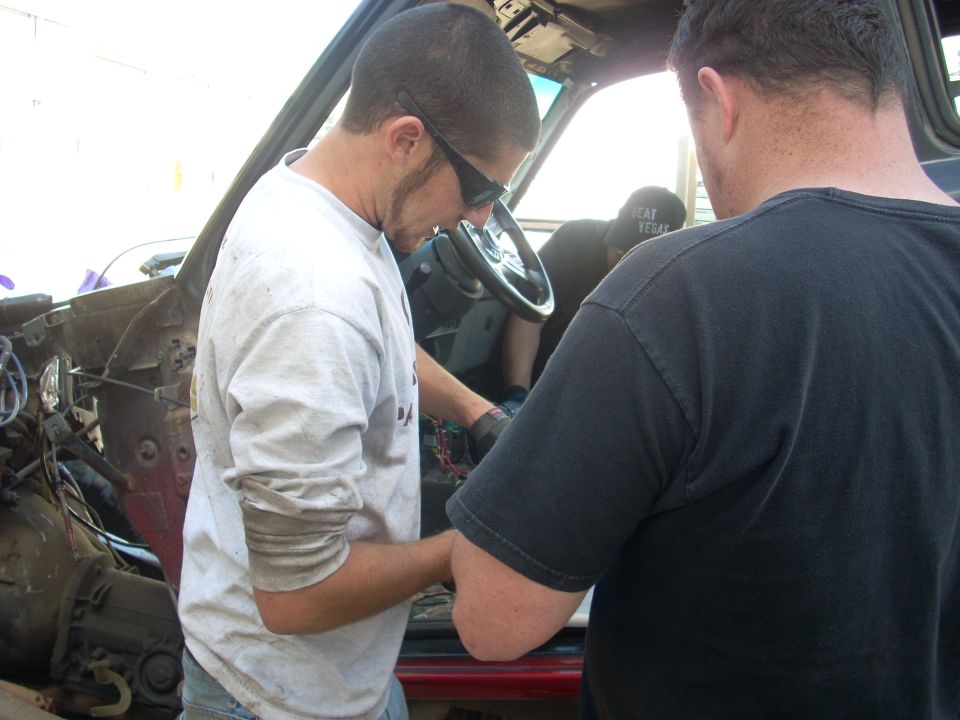 Two guys are working on the wiring inside a vehicle. One is wearing sunglasses and a white shirt, the other is wearing a black shirt